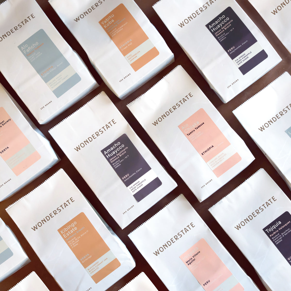 Multiple white Wonderstate coffee bags arranged diagonally on a dark brown surface, each showing a different flavor label in muted blue, peach, orange, pink, and dark purple tones.