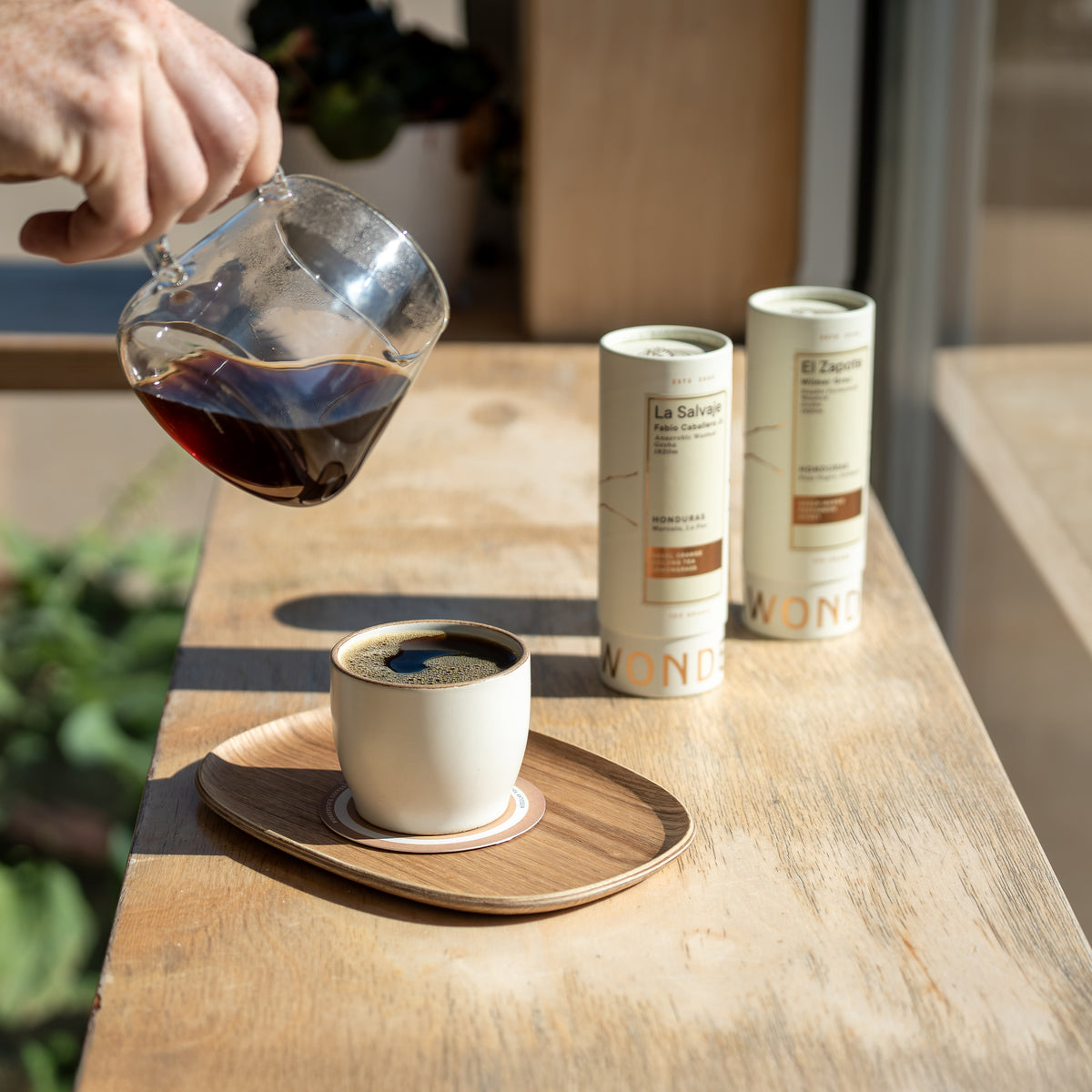 A hand pours dark coffee from a glass pitcher into a small white cup on a wooden saucer, with two cylindrical canisters standing on the table behind. The scene is set on a sunlit wooden table with blurred greenery in the background.