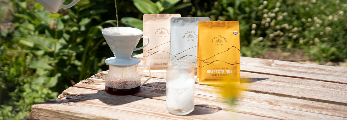 A hand pours hot water from a white pour-over coffee maker into a glass carafe on a wooden table outdoors. Three coffee bags from the brand Mountain Coffee Roasters are standing behind the setup, with a blurred garden background.