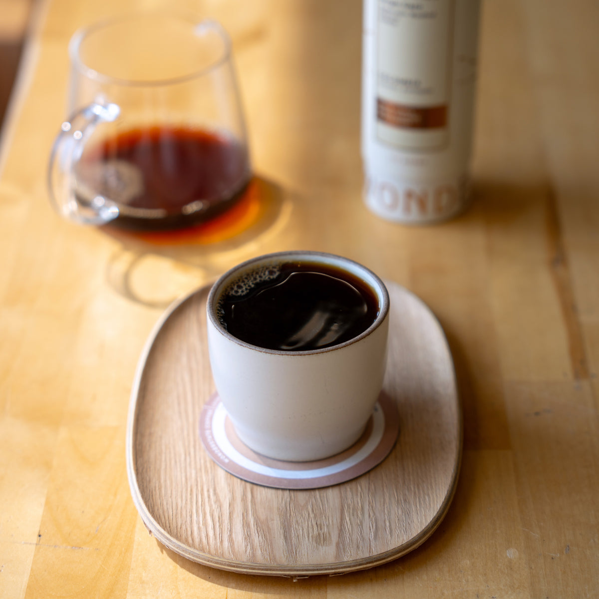A white ceramic cup filled with black coffee sits on a round wooden coaster on a light wood table, with a glass of coffee and a canister in the softly blurred background.