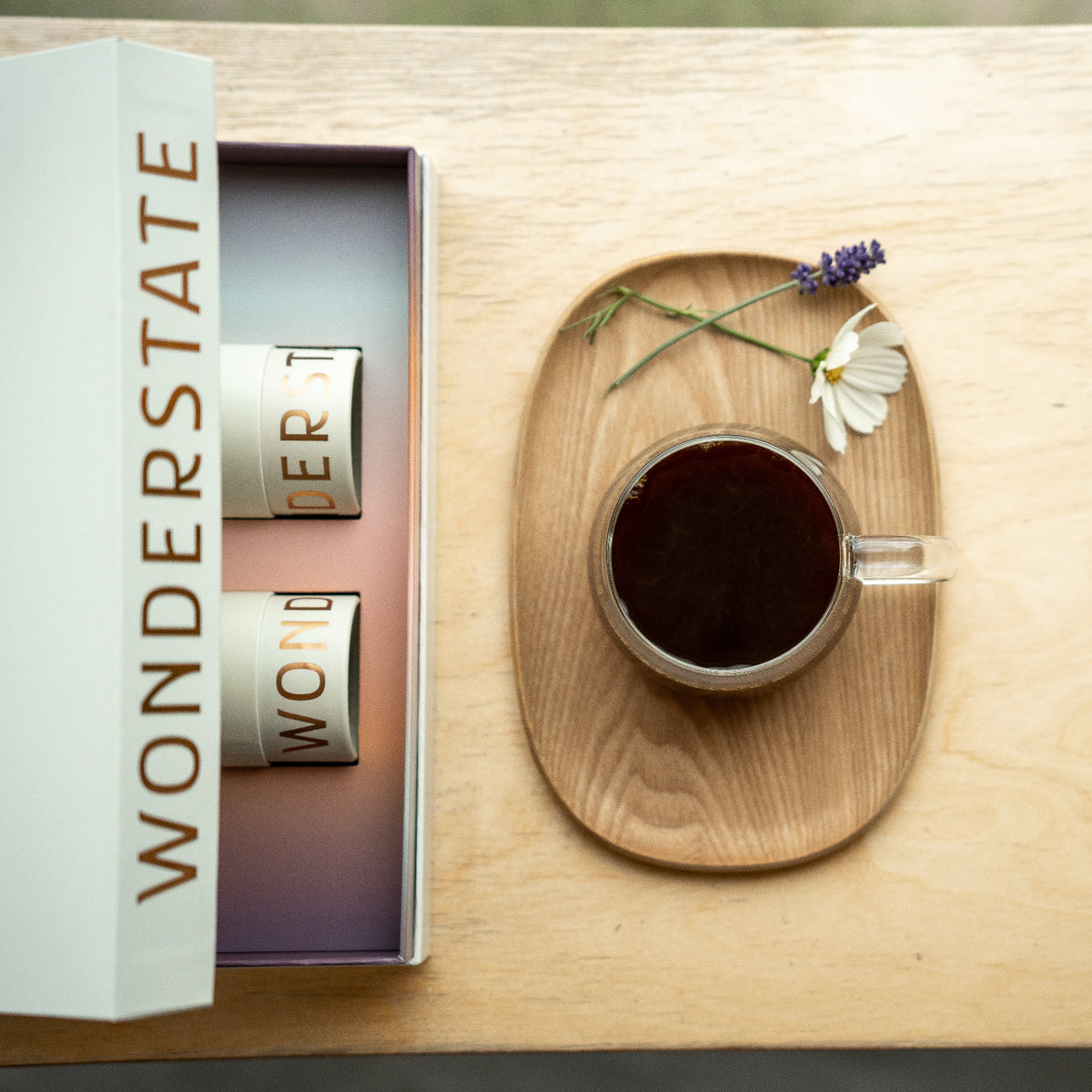 A cup of coffee on a wooden tray with flowers next to a partially open box with two tubes of coffee inside.