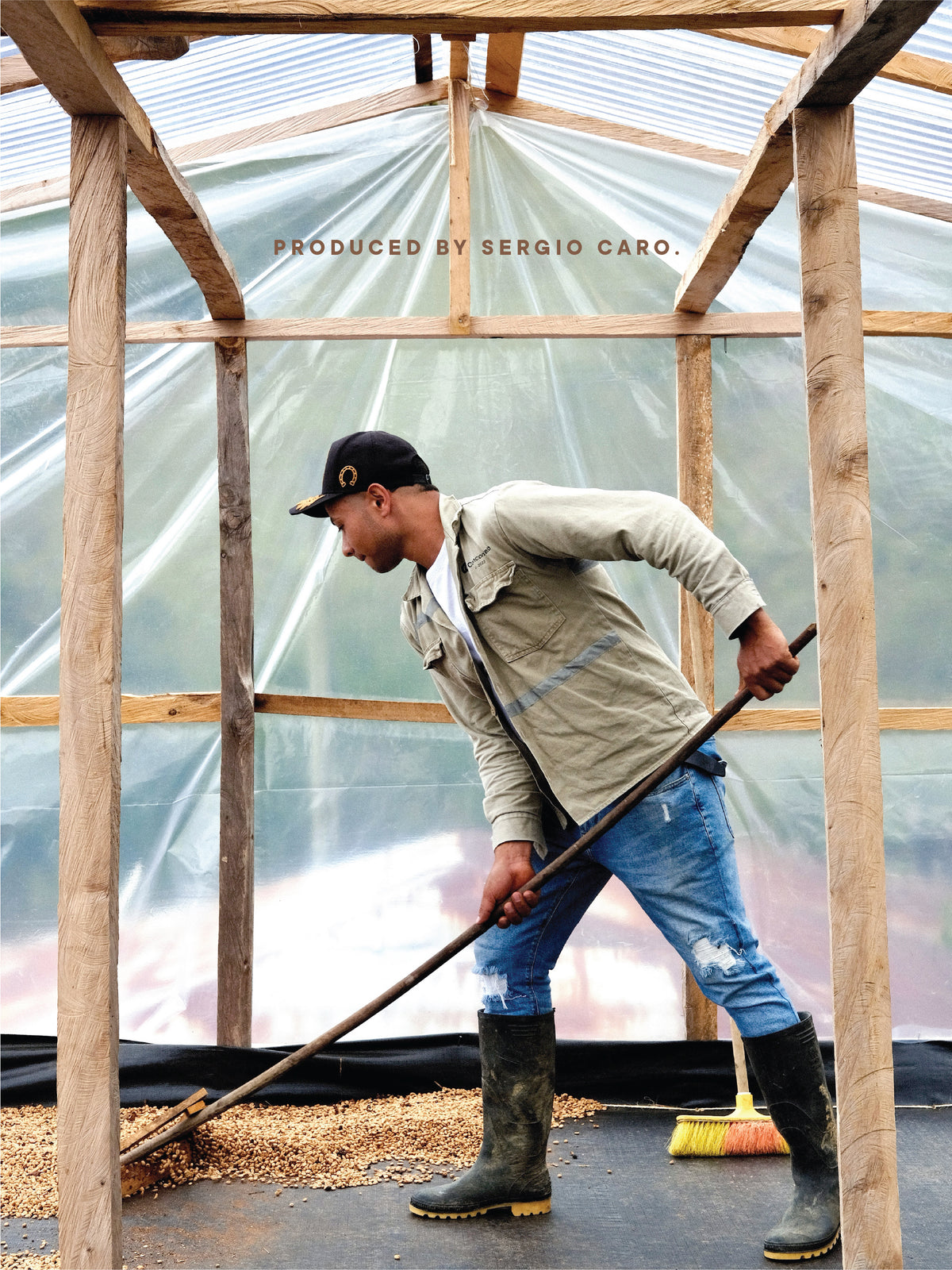 A man in a beige jacket, blue jeans, and dark rubber boots sweeps grain inside a rustic wooden greenhouse structure, with translucent plastic walls behind him. Text at the top reads 'PRODUCED BY SERGIO CARO.