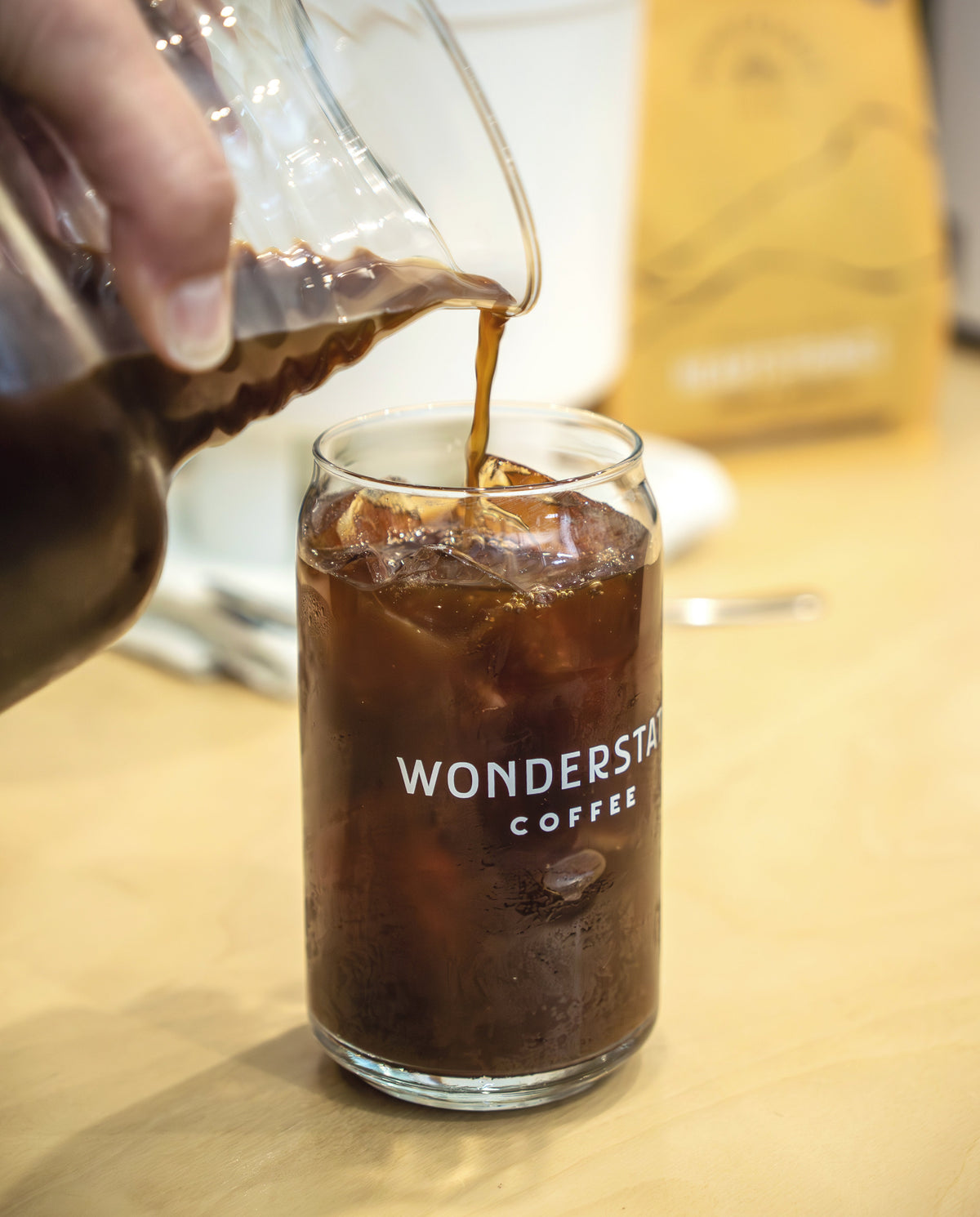 A hand pours dark coffee from a glass pitcher into an iced glass canister labeled 'WONDERSTATE COFFEE' on a light wood table, with a branded coffee bag blurred in the background.
