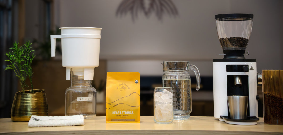 A coffee preparation setup on a countertop, featuring a white manual dripper, a yellow heart-shaped coffee bag, a glass pitcher with water, a glass of ice, and a white coffee grinder with dark coffee beans in its hopper.